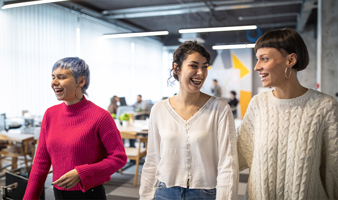 Trois femmes souriantes marchent ensemble dans un bureau moderne.