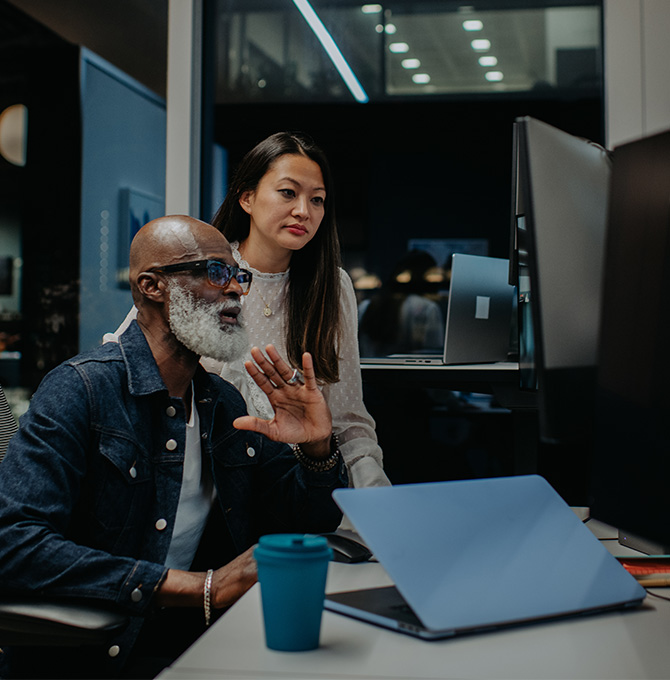 Two technology professionals in a modern office space viewing and discussing information on a computer screen. It is dark outside.