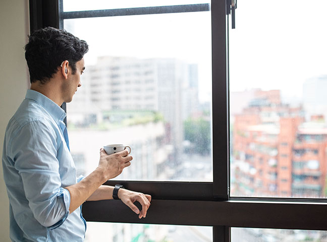 A man with a hearing aid holds a cup of coffee and looks out a window.