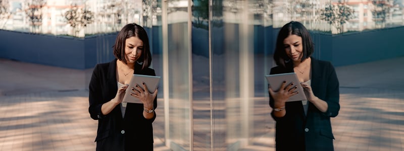 Une femme d’affaires qui tient une tablette est debout à côté d’un édifice à bureaux. Son image se reflète.