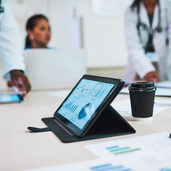 A tablet displaying charts and graphs sits on a table in the foreground. Medical professionals are in the background.