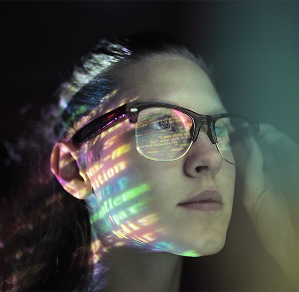 A woman wearing glasses looking off into the distance. Lines of colourful code are lit on her face and glasses.