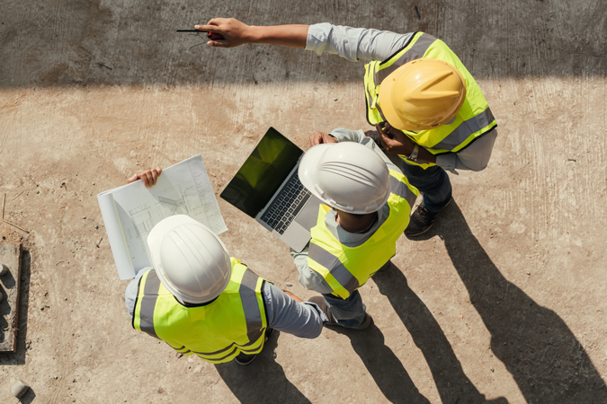 An overhead view of three workers at a construction site wearing hard hats and safety vests. One is holding blueprints, another is holding a laptop, and the other is pointing.