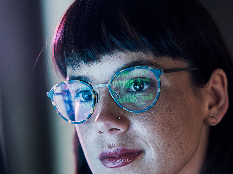 Close-up of a woman's face with data on a screen reflecting in her glasses.