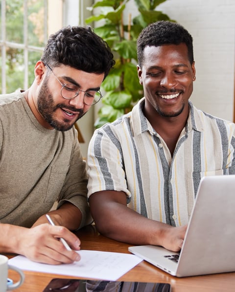 Two men sit at a table in a bright, modern living room, smiling as they work together with a laptop and documents.