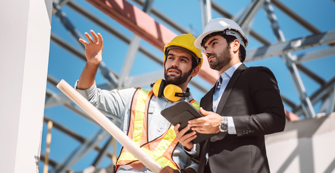 Two men wearing hard hats at a worksite. One is wearing jeans, long-sleeve shirt, and a safety vest while holding rolled up blueprints. The other is wearing a suit and tie and holding a tablet.